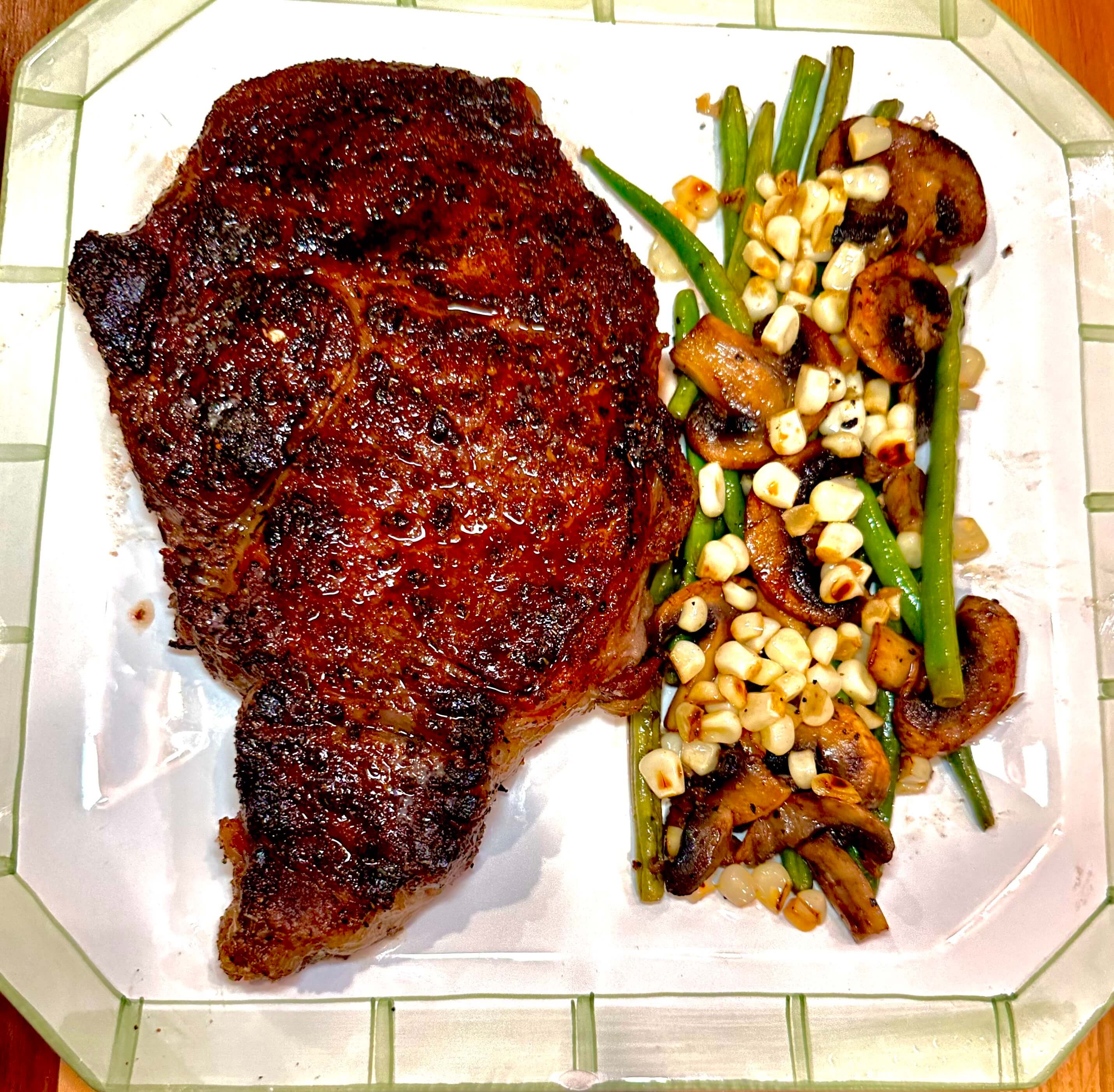 Blackened ribeye steak served with sautéed mushrooms, green beans, and corn on a white plate with green trim.