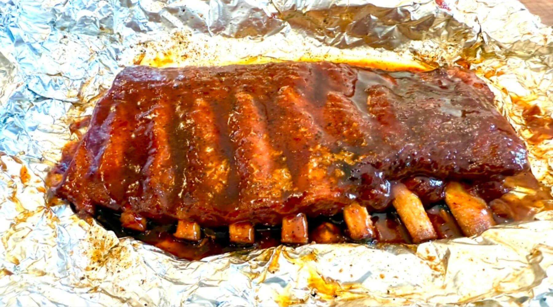 Rack of glazed barbecue ribs resting in a foil boat, with visible sauce and caramelized edges on the meat.