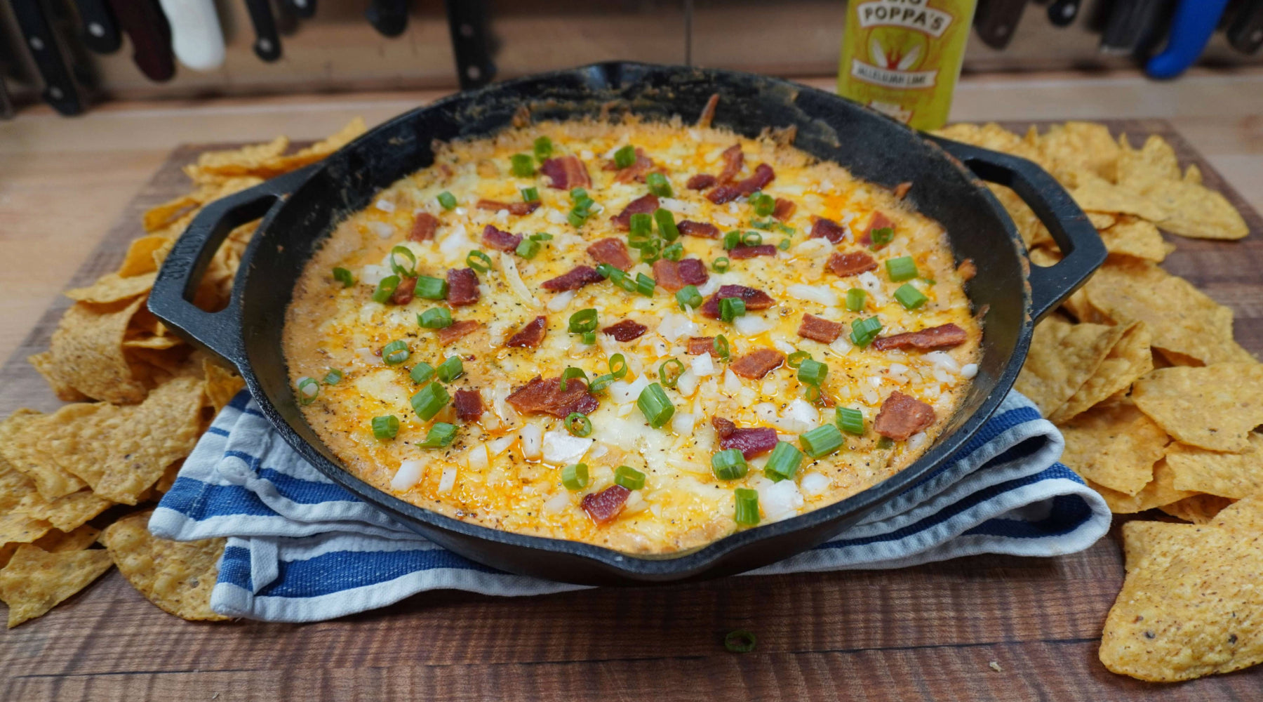 Cast-iron skillet of creamy queso topped with bacon, green onions, and melted cheese, served with tortilla chips.