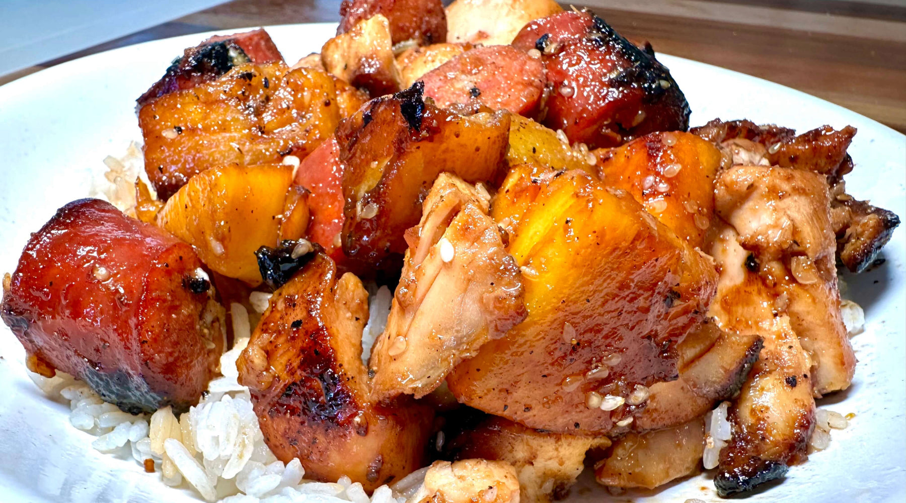Grilled chicken, pineapple, and sausage glazed with sauce, served over white rice on a plate.