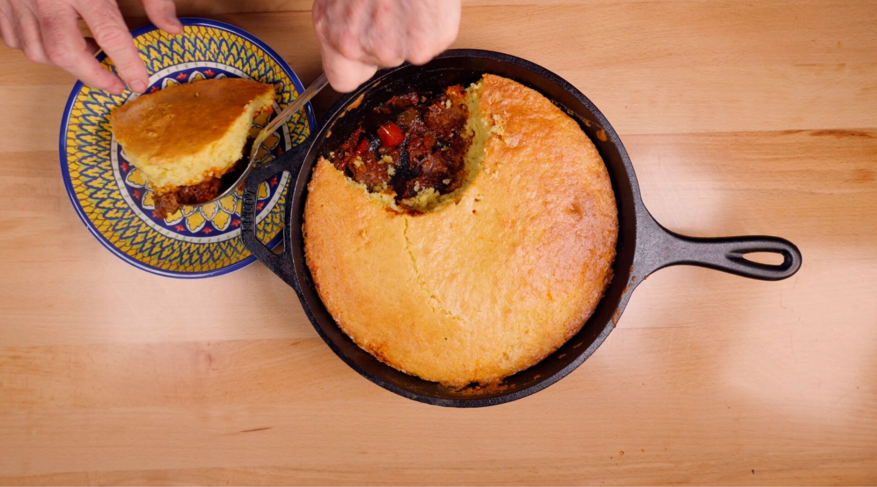 Cast-iron skillet of cornbread topped chili, with a slice being served onto a plate.