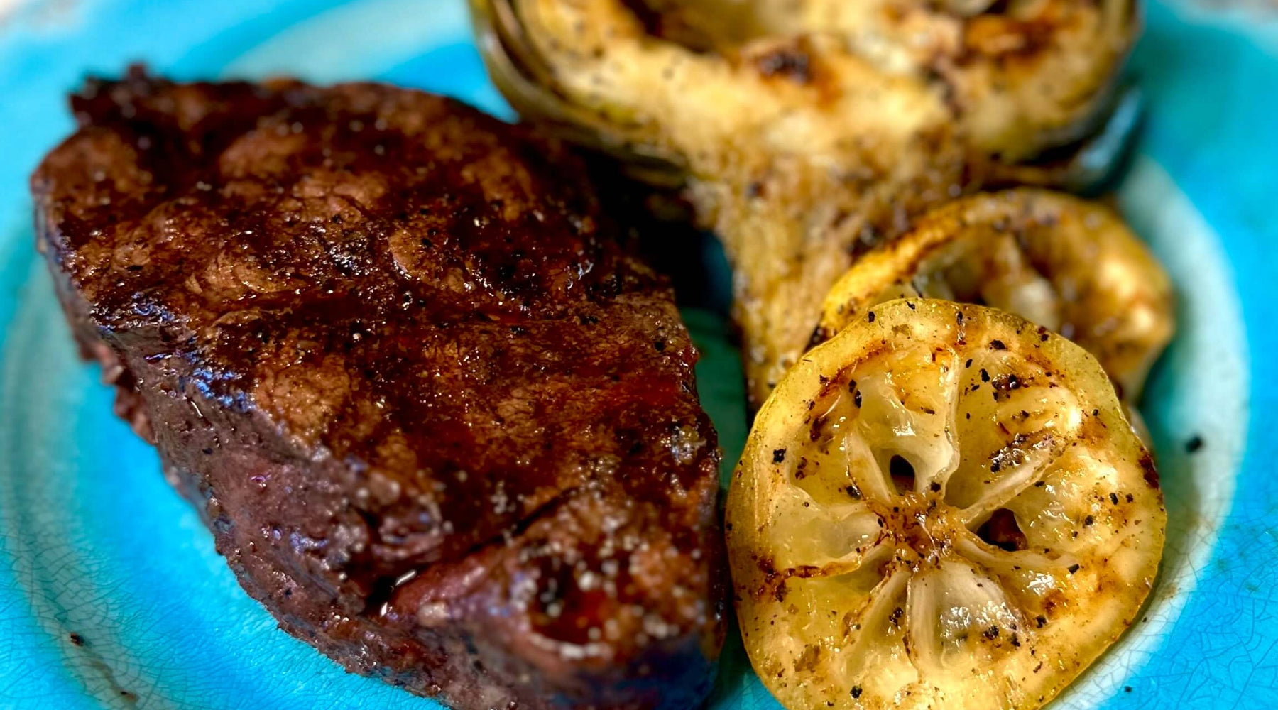 Grilled steak served with charred artichoke halves and seared lemon slices on a rustic blue plate.