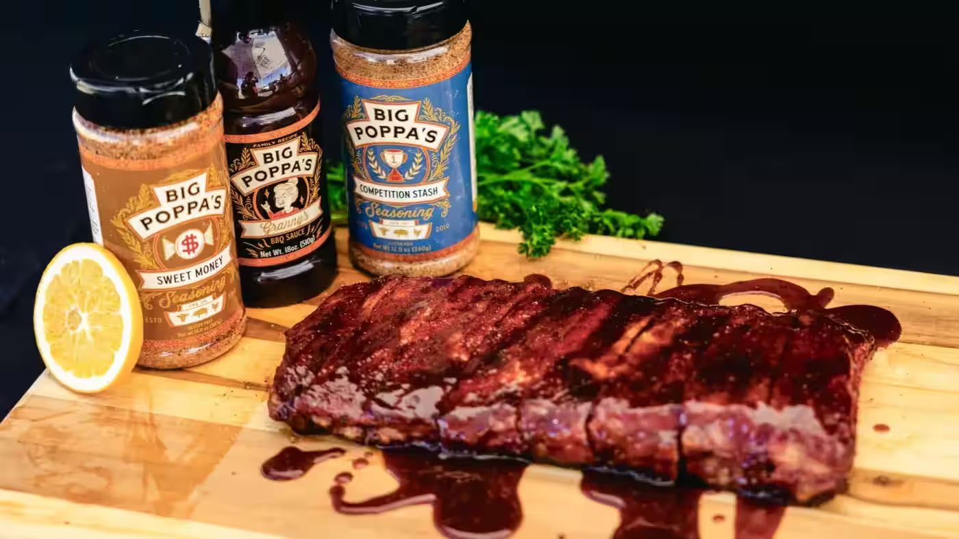 Grilled ribs on a cutting board with Big Poppa's barbecue sauce bottles and ingredients.
