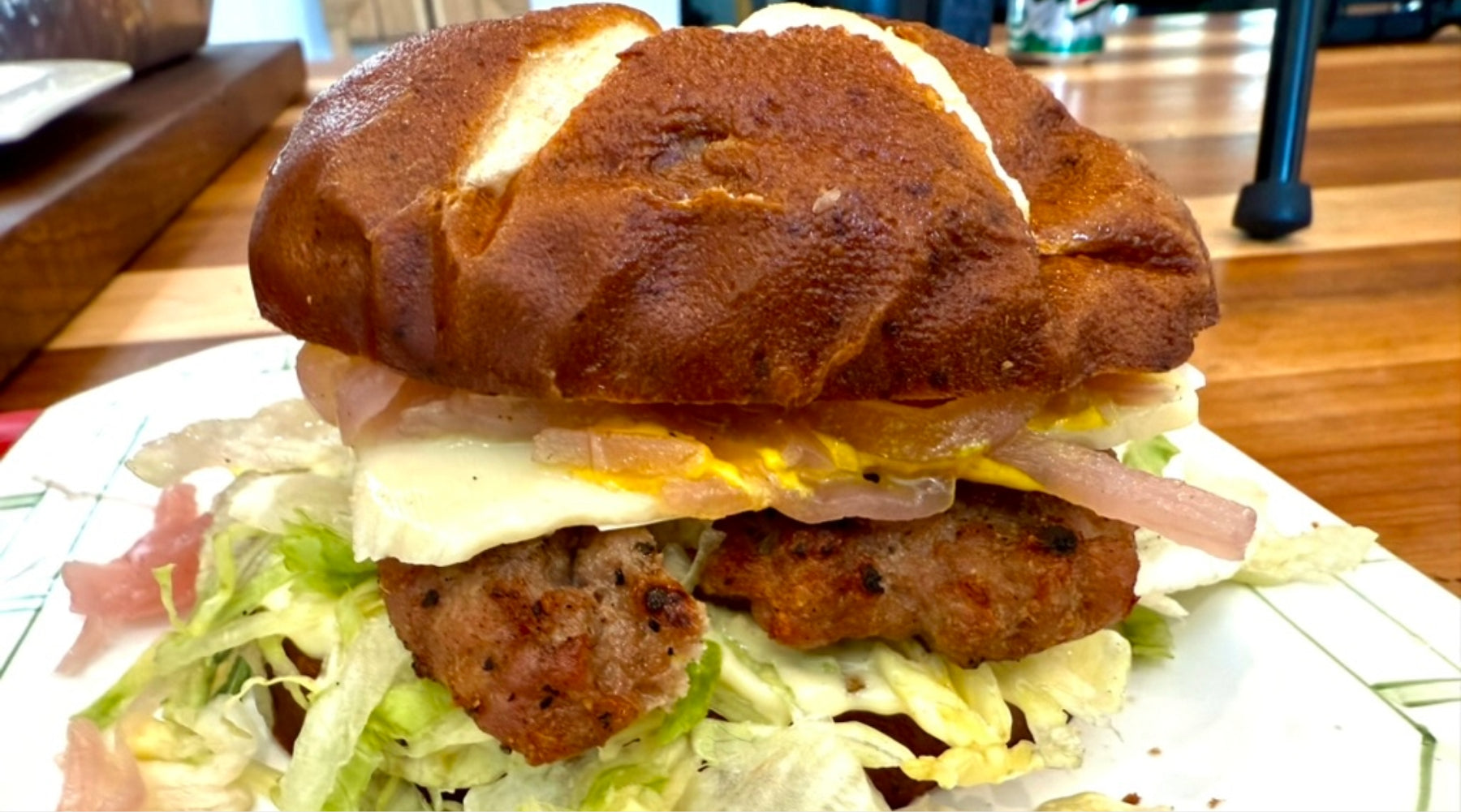 A sandwich with sausage patties, shredded lettuce, fried egg, and onions on a pretzel bun, served on a white plate.