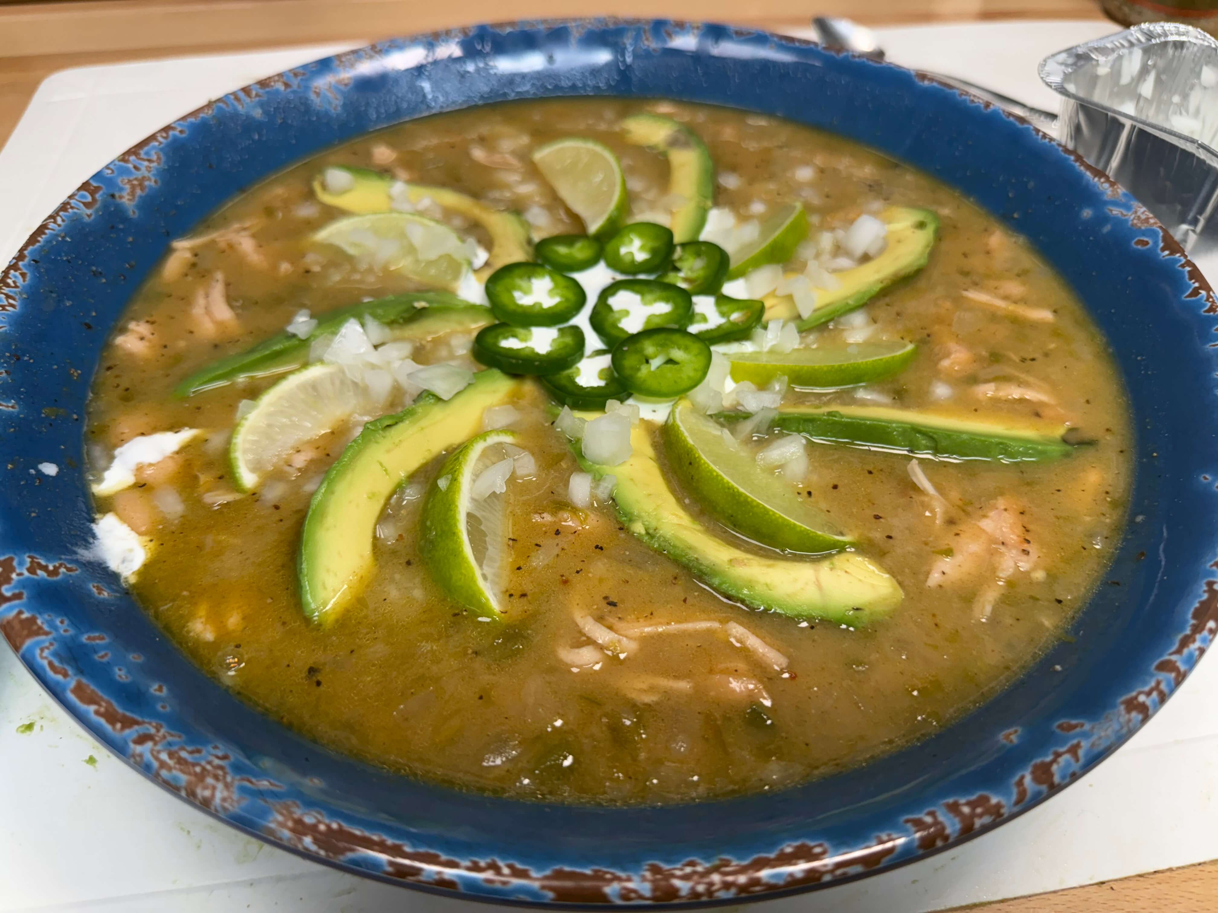 Bowl of chicken soup topped with avocado slices, lime wedges, jalapeños, and diced onions in a green broth.