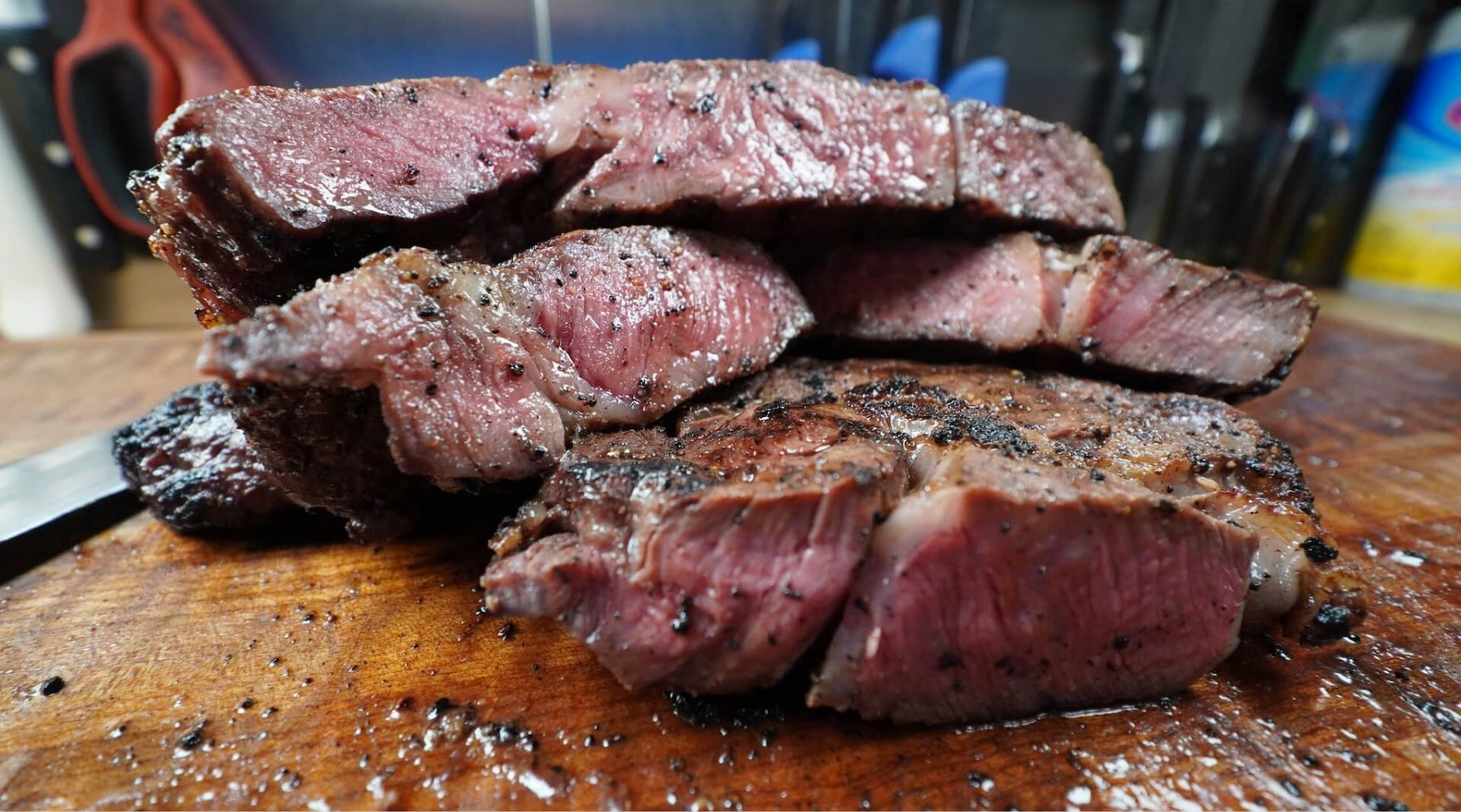 Sliced medium-rare steak with peppered crust, resting on a wooden cutting board with juices visible.
