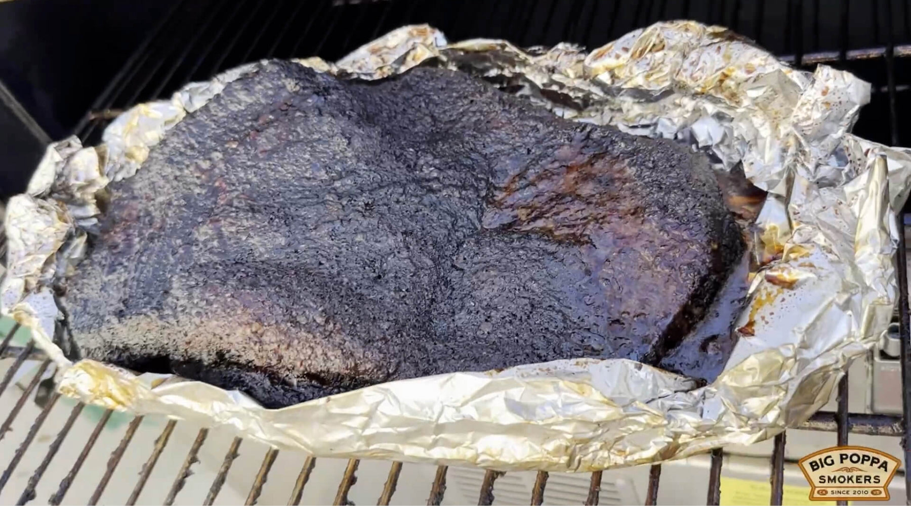 Large smoked brisket with dark bark resting in foil on a grill grate, juices pooling around the meat.