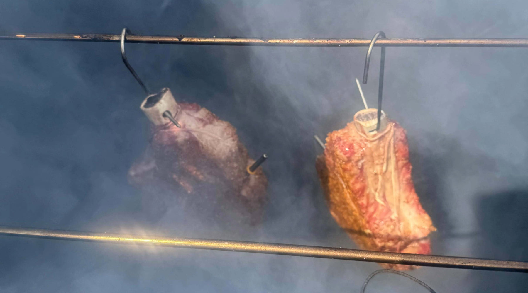 Two cuts of meat hanging on hooks inside a smoker, surrounded by smoke as they cook.