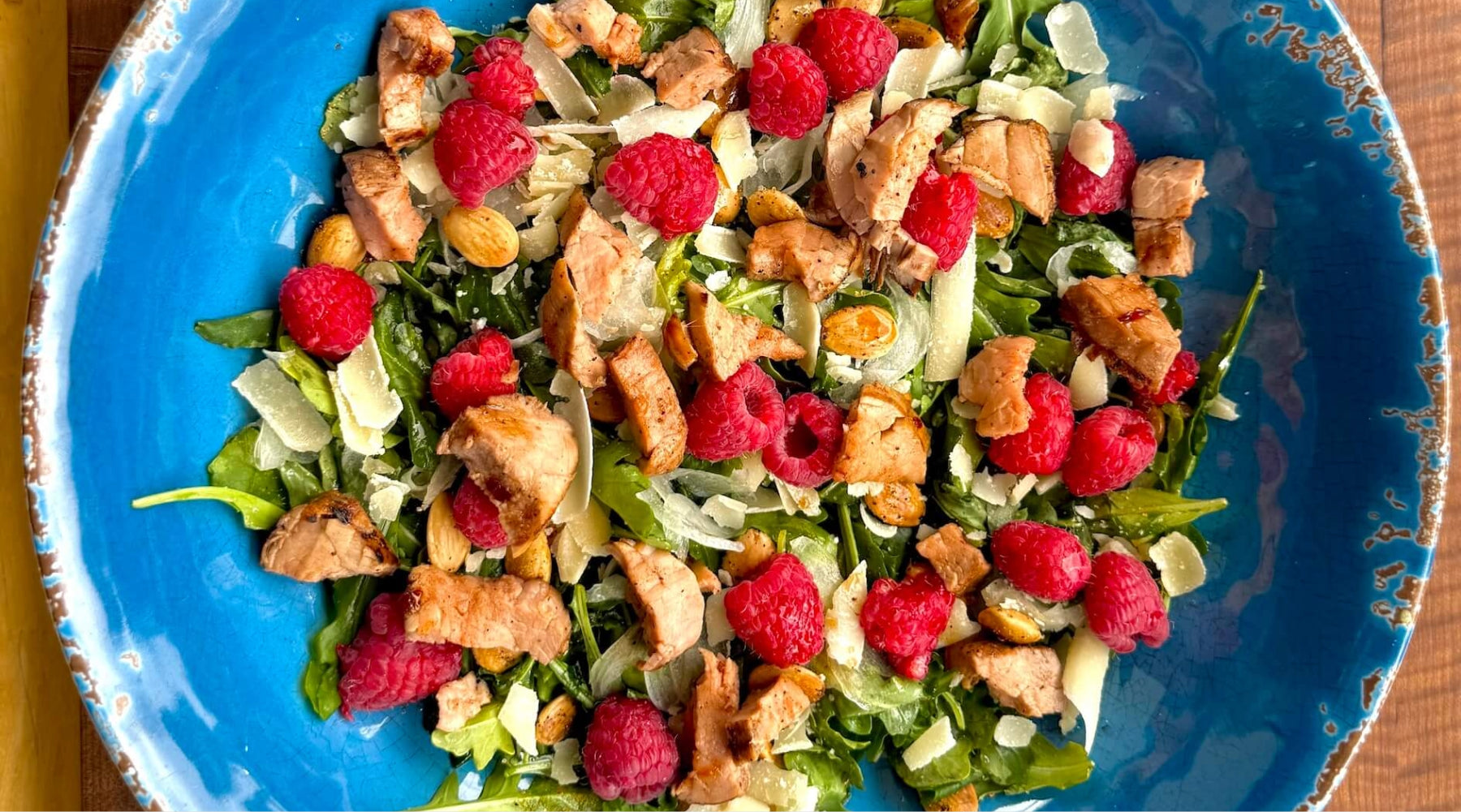 Salad with grilled chicken, raspberries, almonds, and shaved cheese on mixed greens in a blue bowl.
