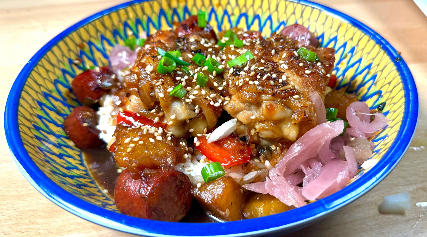Bowl of glazed chicken with sausage, peppers, and rice, topped with sesame seeds, green onions, and pickled onions.