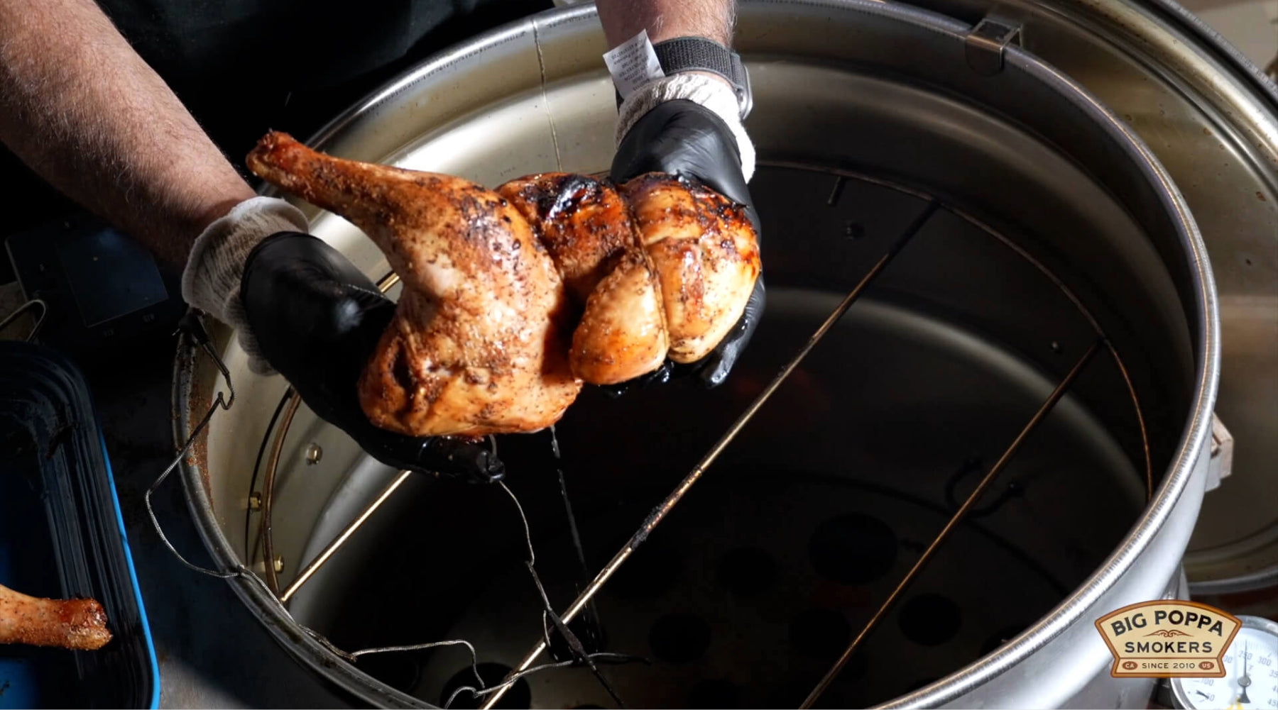 Gloved hands lifting a whole smoked chicken from a drum smoker, juices dripping from the browned skin.