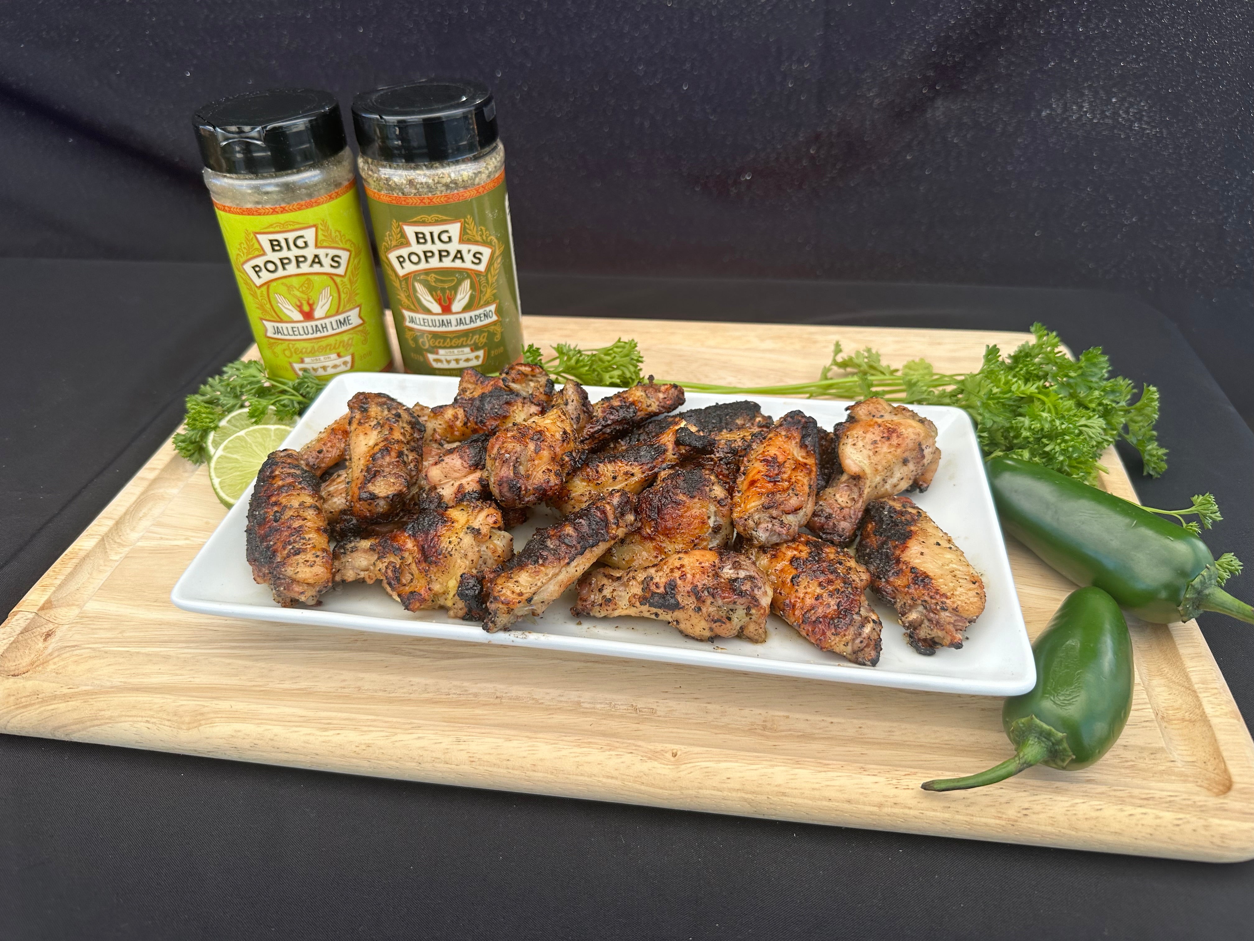 A white platter of grilled chicken wings, garnished with fresh parsley and jalapeños, with jars of Jallelujah seasonings nearby.